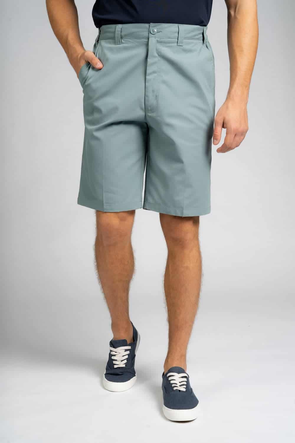 Front view of a person wearing sage green knee-length shorts with a navy polo shirt, one hand in the pocket, paired with blue lace-up sneakers.